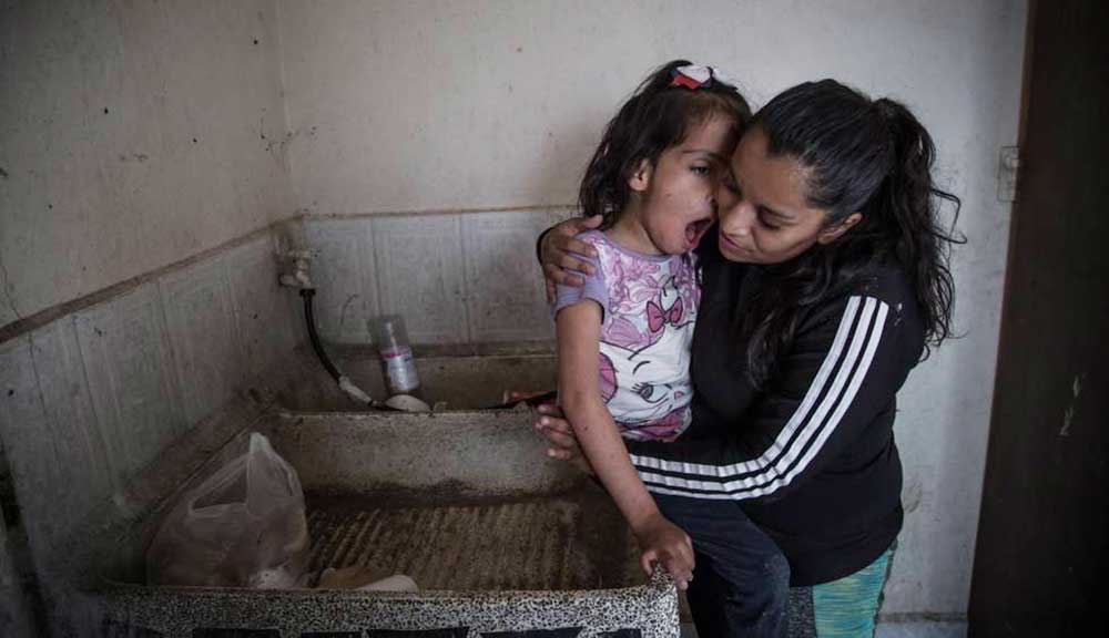 Foto de una mujer joven tomando en brazos a su hija con discapacidad dentro de una cocina que se nota muy precaria.